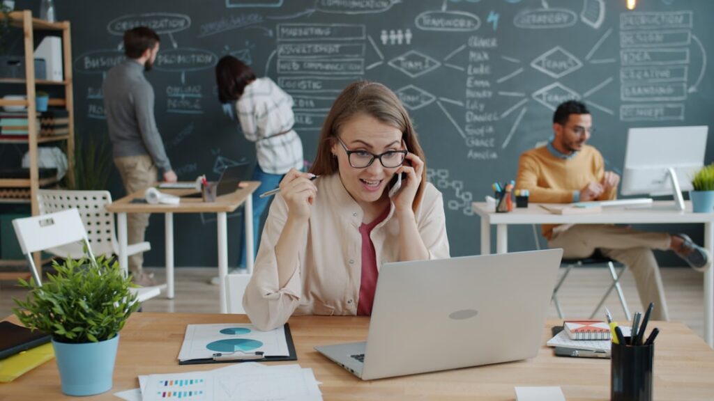 top digital marketing trends — Happy girl blonde is talking on mobile phone showing thumbs-up at desk in coworking workplace while people talking and working with chalkboard in background.