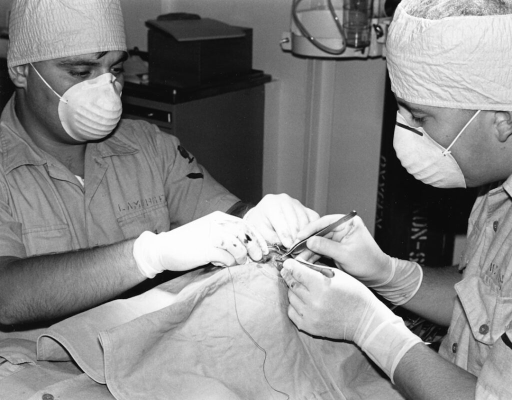 15-3001-0172 HM3 Michael Lamorgese (left) and HM3 David McGinnis suture a minor head laceration. [Corpsmen][Patient][Surgery] "With Iowa at Sea" by J.K. Herman Published in Navy Medicine Magazine Sept-Oct 1986